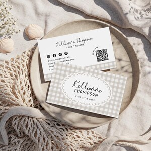 May include: Two business cards with the name "Kellianne Thompson" printed on them. One card has a gingham pattern and the other has social media icons. The cards are on a neutral-colored plate with seashells and flowers.