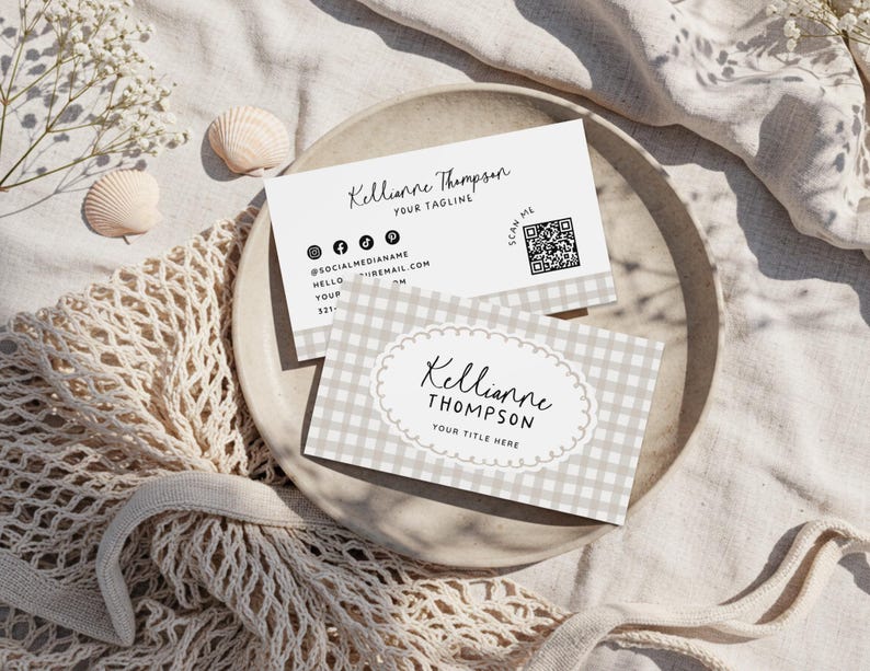 May include: Two business cards with the name "Kellianne Thompson" printed on them. One card has a gingham pattern and the other has social media icons. The cards are on a neutral-colored plate with seashells and flowers.