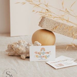Puede incluir: Una tarjeta de visita con el texto "Rainbow Eclectic Baby Boutique" en una fuente colorida y caprichosa. La tarjeta est&aacute; sobre una superficie marr&oacute;n claro con un peque&ntilde;o trozo de coral y una esfera dorada.
