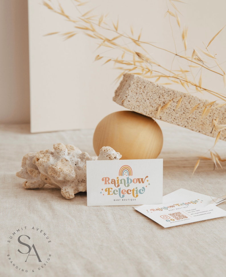 Puede incluir: Una tarjeta de visita con el texto "Rainbow Eclectic Baby Boutique" en una fuente colorida y caprichosa. La tarjeta est&aacute; sobre una superficie marr&oacute;n claro con un peque&ntilde;o trozo de coral y una esfera dorada.