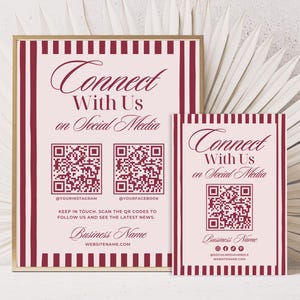 May include: Two framed signs with a pink background and burgundy stripes. The signs read "Connect With Us on Social Media" and include QR codes for Instagram and Facebook. The business name and website are also displayed.