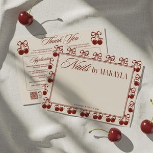 May include: Two cream-colored business cards with burgundy cherry and bow illustrations. The cards read "Nails by MAKAYLA" and "Thank You." Fresh cherries with stems are scattered around the cards on a white fabric surface.