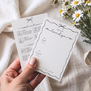Könnte beinhalten: Zwei weiße Karten mit schwarzem Text und Illustrationen werden in einer Hand gehalten. Auf einer Karte steht "nails by londyn" mit einem Schleifendesign. Die andere Karte enthält Anwendungs- und Entfernungsanweisungen. Gänseblümchen sind im Hintergrund.