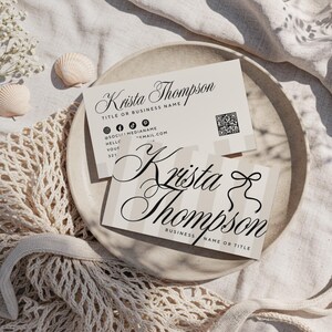 May include: Two business cards with the name "Krista Thompson" in elegant script are displayed on a neutral-toned plate. One card has a striped design, and both include contact information and a QR code. The cards are surrounded by seashells and a net bag.