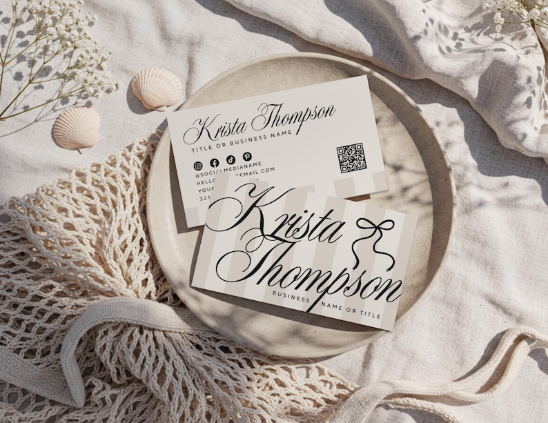 May include: Two business cards with the name "Krista Thompson" in elegant script are displayed on a neutral-toned plate. One card has a striped design, and both include contact information and a QR code. The cards are surrounded by seashells and a net bag.