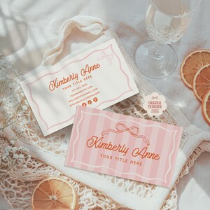 Op de afbeelding: Twee visitekaartjes met de naam "Kimberly Anne" in een sierlijk lettertype. Eén kaart is wit met een roze rand en strik, de andere is roze met verticale strepen. Gedroogde sinaasappelschijfjes en een wijnglas zijn ook zichtbaar.