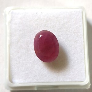 Puede incluir: Una piedra preciosa de rubí de forma ovalada y facetada con un tono rojo rosáceo intenso. La gema se muestra centrada sobre un inserto de espuma blanca cuadrada dentro de un pequeño recipiente de almacenamiento de plástico blanco.