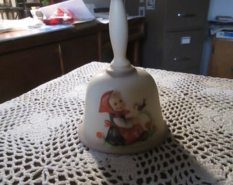 May include: A white ceramic bell with a handle and a painted image of a girl in a red dress holding a bird. The bell has a brown base and is decorated with orange flowers.