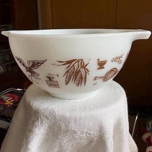 May include: White Pyrex mixing bowl with brown illustrations of an eagle, corn, and other kitchen-themed designs. The bowl has two handles and is sitting on a white cloth.