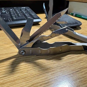May include: A silver multi-tool with several extended implements, including a serrated blade, a flat-head screwdriver, and a metal ruler with markings up to 2 inches. The tool features a metallic finish and a hinged design with multiple folding parts.