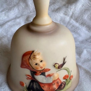 May include: A ceramic bell with a cream-colored body and a handle. The bell features a painted scene of a girl in a red hood and dress, with a bird perched nearby. The bell has a brown base and is likely a decorative item.