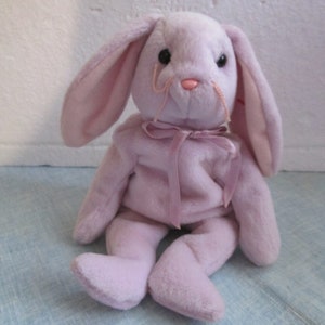 May include: A plush lavender bunny with long ears and a pink bow. The bunny is sitting with its legs crossed.