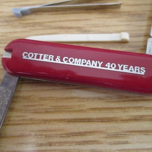 May include: A red pocket knife with a silver blade and a white handle. The knife has the text "COTTER & COMPANY 40 YEARS" printed on the side.