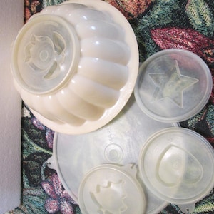 May include: A set of four vintage clear plastic jelly molds with different designs. The molds are in various shapes, including a star, a bell, a heart, and a flower. The molds are all in good condition and have a slightly yellowed tint.