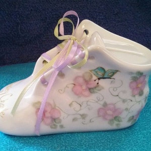May include: A white porcelain baby shoe with a floral design and a butterfly. The shoe has a lavender and yellow ribbon tied around it. The shoe says "Baby's Choice" on the side.