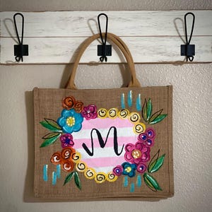 以下が含まれることがあります： 花柄と文字「M」が入った、ベージュ色のジュートトートバッグ。 花柄には青、ピンク、オレンジ、黄色の花が含まれています。 バッグにはベージュ色のハンドルが付いており、黒いフックが付いた白い木板に掛けられています。