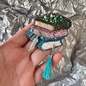 May include: A handcrafted brooch in the shape of stacked books, embellished with beads in shades of green, pink, and teal. A silver feather charm and a teal tassel dangle from the bottom. The brooch is a unique accessory.