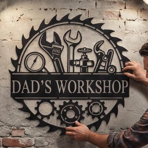Pode incluir: Placa de metal preta com o texto "DAD'S WORKSHOP" e silhuetas de ferramentas. A placa tem a forma de uma lâmina de serra e engrenagens na parte inferior. Está sendo montada em uma parede de tijolos.