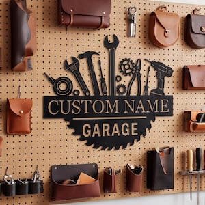 May include: A black metal wall sign featuring a circular saw blade design with silhouettes of tools including a wrench, hammer, screwdriver, and drill. The text reads CUSTOM NAME and GARAGE. It is mounted on a tan pegboard with leather pouches.
