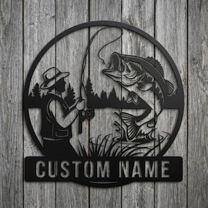 May include: Black metal wall art depicting a fisherman reeling in a large fish. The design includes a fisherman silhouette, a leaping fish, and the text "CUSTOM NAME" on a rectangular plaque. The artwork is set against a wooden background.