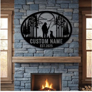 May include: A black oval metal wall sign featuring a silhouette of a hunter with a rifle and a dog standing in a forest under a large moon. The text CUSTOM NAME and EST.2025 is cut out at the bottom of the design, creating a decorative display piece.