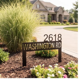 Pode incluir: Uma placa de endereço de metal preto com o número 2618 e o texto WASHINGTON RD em estilo recortado. A placa é montada em duas estacas e posicionada em um canteiro de jardim com cobertura morta, plantas verdes e pequenas flores coloridas.