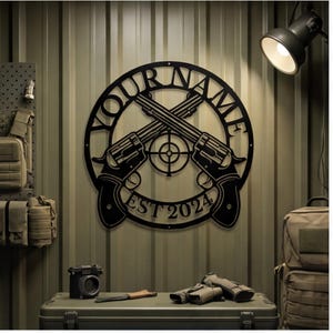 May include: A circular black metal wall sign featuring two crossed revolvers and a central target symbol. The text "YOUR NAME" is arched across the top, and "EST 2024" is positioned along the bottom curve of the circular frame design.