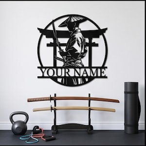Op de afbeelding: Een cirkelvormig wandkunstwerk van zwart metaal met het silhouet van een samoerai die een conische hoed draagt en een katana vasthoudt voor een torii-poort. Het ontwerp bevat een horizontale balk met de tekst YOUR NAME in een schreeflettertype.