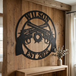 Op de afbeelding: Zwarte metalen wandkunst met gekruiste revolvers in een cirkelvormig frame. Het woord "WILSON" staat bovenaan, met "EST 2025" eronder. De kunst is bevestigd aan een houten muur.
