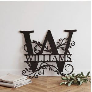 Pode incluir: Uma placa de parede em metal preto com uma letra maiúscula W grande e o nome WILLIAMS recortado no centro. O design inclui padrões intrincados de vinhas e folhas entrelaçadas integrados na estrutura da letra, criando uma peça decorativa.