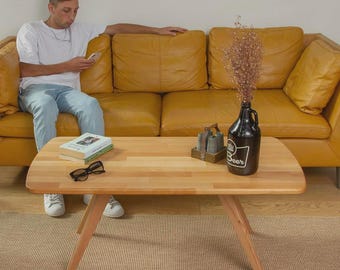 May include: A light-coloured wooden coffee table with angled legs. A black bottle with the word "Beer" is on the table, along with a small decorative item and a pair of sunglasses. A yellow leather sofa is in the background.