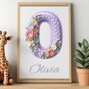 May include: A framed artwork featuring a lavender-colored, crocheted number zero adorned with colorful floral accents. The name "Olivia" is written in a matching lavender script below the number. The artwork is displayed in a wooden frame.