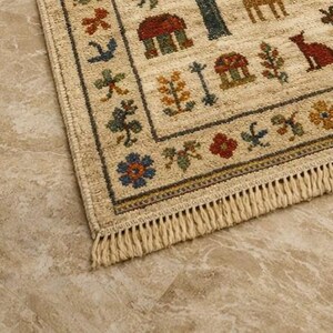 Pu&ograve; includere: Tappeto rettangolare beige con bordo e frange dettagliati. Il tappeto presenta un motivo ripetuto di case, alberi, animali e motivi floreali nei toni del rosso, blu, verde e giallo. Il tappeto &egrave; posato su una superficie marrone chiaro.