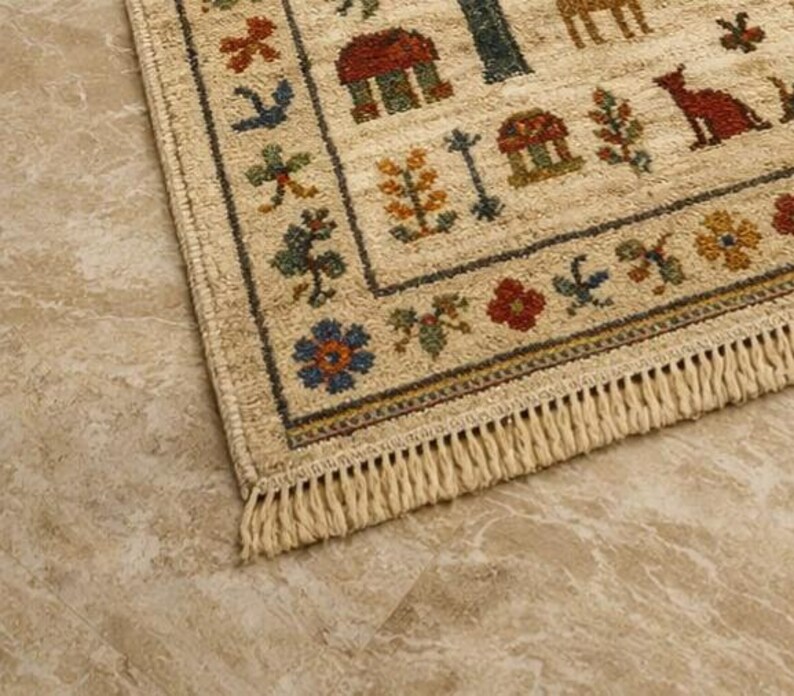 Pu&ograve; includere: Tappeto rettangolare beige con bordo e frange dettagliati. Il tappeto presenta un motivo ripetuto di case, alberi, animali e motivi floreali nei toni del rosso, blu, verde e giallo. Il tappeto &egrave; posato su una superficie marrone chiaro.