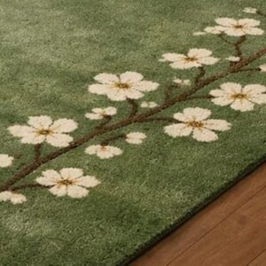 Puede incluir: Alfombra verde con dise&ntilde;o floral. La alfombra presenta una rama con flores blancas y centros amarillos. La alfombra est&aacute; sobre un suelo de madera. La alfombra es de un material suave y afelpado.