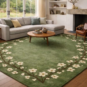 Puede incluir: Una alfombra verde con un dise&ntilde;o floral. La alfombra presenta un borde de flores blancas y ramas marrones sobre un fondo verde liso. La alfombra est&aacute; en un sal&oacute;n con un sof&aacute; seccional gris y una mesa de centro de madera.