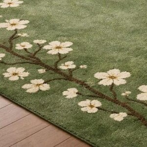 Puede incluir: Alfombra verde con dise&ntilde;o floral. La alfombra presenta una rama marr&oacute;n con flores blancas. La alfombra es de color verde suave y tiene una apariencia texturizada. Est&aacute; colocada sobre un suelo de madera.