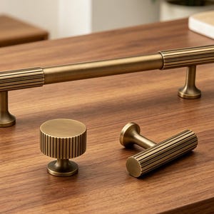 Puede incluir: Un conjunto de herrajes de latón para armarios, que incluye un tirador largo de barra, un pomo redondo y un tirador más pequeño. Los herrajes presentan un diseño acanalado y se exhiben sobre una superficie de madera.