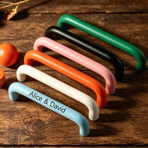 May include: A collection of colorful cabinet handles in green, black, pink, orange, white, and light blue. The light blue handle has the text "Alice & David". Also included are two orange cabinet knobs. The handles are arranged on a wooden surface.