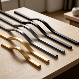 Puede incluir: Una colección de tiradores de armario modernos en varios acabados: dorado, plateado y negro. Los tiradores tienen un diseño curvo en forma de onda y están dispuestos sobre una superficie de madera clara, mostrando su estética elegante.