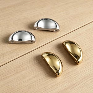 Può includere: Quattro maniglie per mobili a forma di mezzaluna. Due sono argentate lucide e due dorate. Ogni maniglia ha due fori per il montaggio. Le maniglie sono disposte su una superficie di legno chiaro.