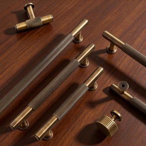 Może przedstawiać: Asortyment antycznych mosiężnych okuć do szafek, w tym uchwytów i gałek. Uchwyty mają żebrowaną część środkową i są zamontowane na ciemnej drewnianej powierzchni. Okucia są w różnych rozmiarach i orientacjach.