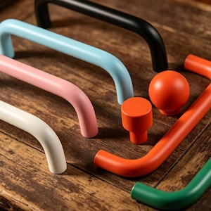 Op de afbeelding: Een verzameling kleurrijke kastbeslagen, waaronder gebogen handgrepen in zwart, lichtblauw, roze en wit. Er zijn ook oranje knoppen en een groene handgreep, allemaal gerangschikt op een houten oppervlak.