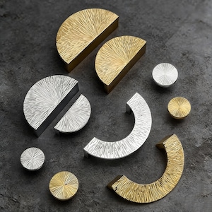 Puede incluir: Una variedad de herrajes decorativos para armarios en tonos dorados y plateados. Los herrajes incluyen tiradores semicirculares, pomos redondos y manijas curvas, todos con un diseño texturizado en forma de sol. Sobre una superficie gris oscura.