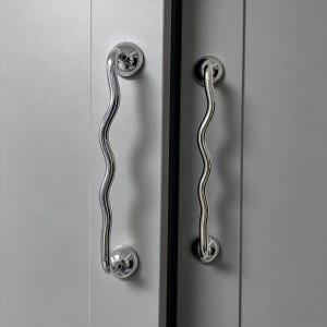 Könnte beinhalten: Zwei verchromte Schrankgriffe mit wellenförmigem, serpentinenartigem Design. Jeder Griff ist auf einer kreisförmigen Basis montiert. Die Griffe sind an einer hellgrauen Schranktür befestigt.