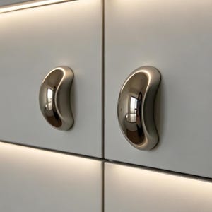 May include: Close-up of two polished, silver-toned cabinet pulls with a smooth, curved design. The pulls are mounted on a light gray cabinet door, reflecting the surrounding environment. The design is modern and minimalist.