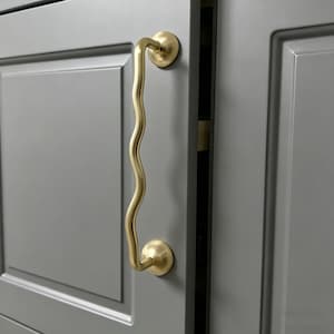Może przedstawiać: Złoty uchwyt do szafki o falistym, pionowym wzorze. Uchwyt jest przymocowany do szarych drzwi szafki z wpuszczanym panelem. Okucia mają okrągłą podstawę na każdym końcu.