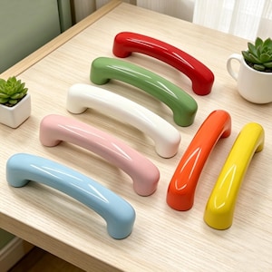 Puede incluir: Primer plano de siete tiradores de armario de colores brillantes, colocados sobre una superficie de madera clara. Los tiradores son curvos y vienen en rojo, verde, blanco, rosa, naranja, amarillo y azul. En el fondo se ve una pequeña planta suculenta en una maceta blanca.