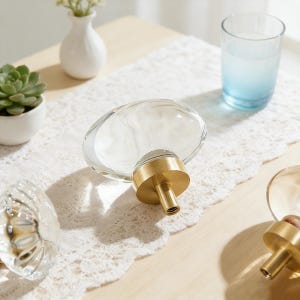 May include: Clear glass oval cabinet knobs with brass bases are displayed on a table. The knobs are partially transparent, allowing light to pass through. A small succulent in a white pot and a glass of water are also visible.