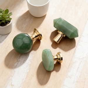 Può includere: Tre pomelli per mobili in giada verde con basi color oro sono disposti su una superficie di legno chiaro. Un pomello è rotondo, uno rettangolare e uno ovale. Una piccola pianta grassa in vaso e una tazza bianca sono sullo sfondo.